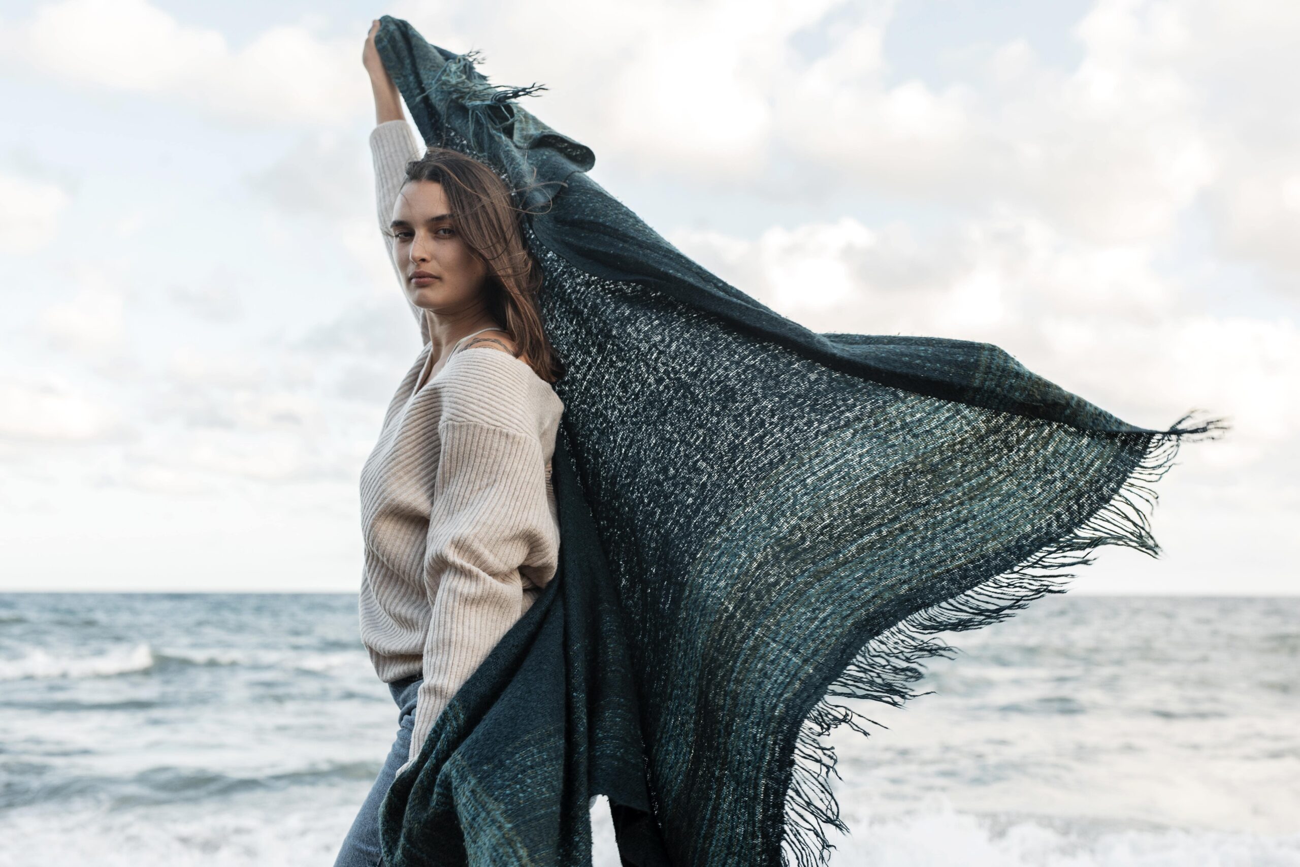 Elegant woman wearing a beige sweater holding a flowing luxury shawl by the seaside on a breezy day.