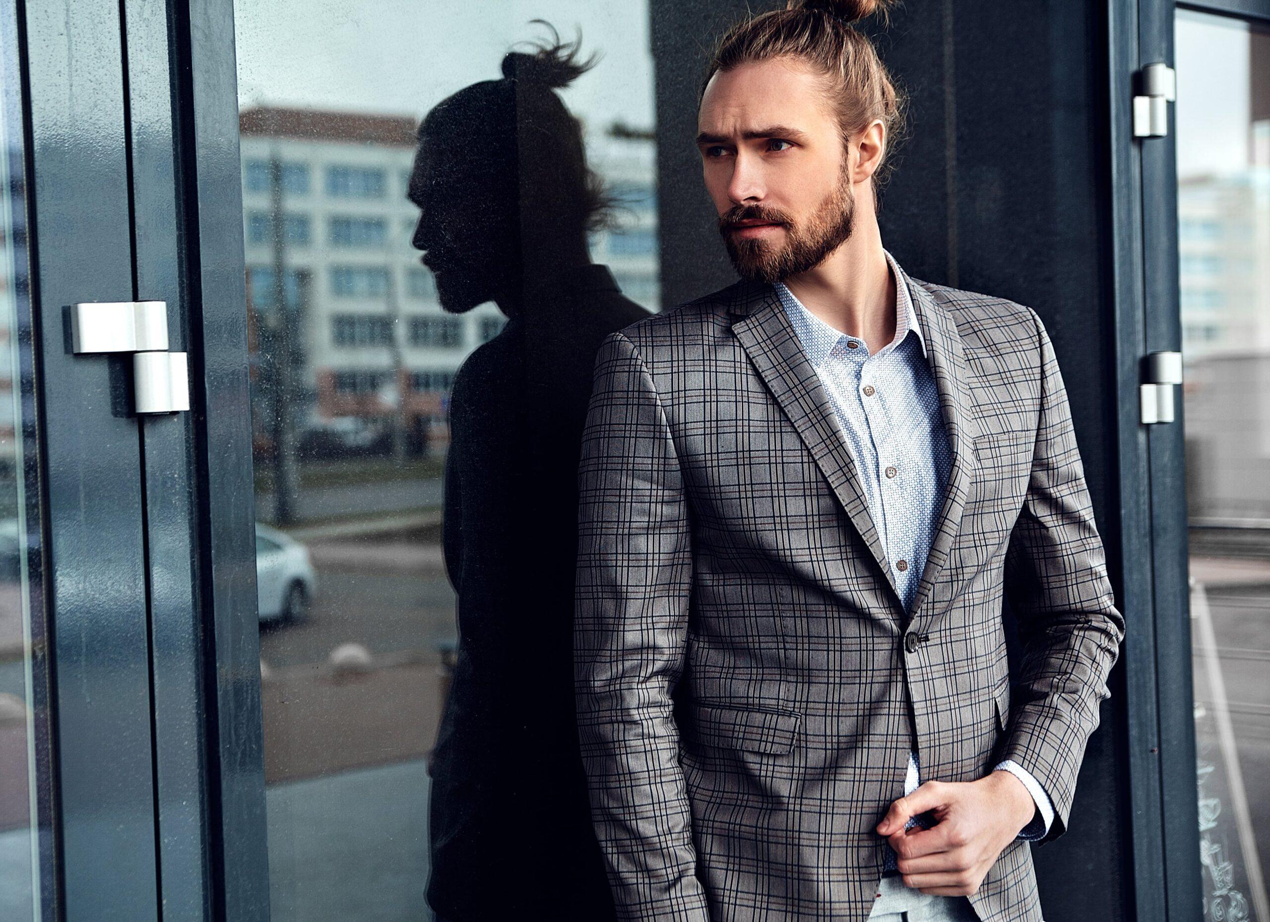 A stylish man wearing a checked blazer and a patterned shirt standing by a glass door with his reflection visible. His hair is tied in a bun, and he exudes a modern, sophisticated look.
